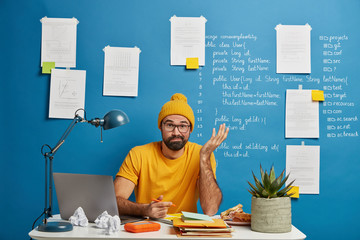 Concerned uncertain male student solves difficult problem while working in studyroom, writes down on iformation, makes essay, has lack of idea, wears yellow clothes, uses internet on laptop computer