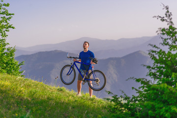 Fototapeta premium Tired cyclist is wiping his sweat off his face while pushing his bicycle uphill on a dirt road in a mountain.