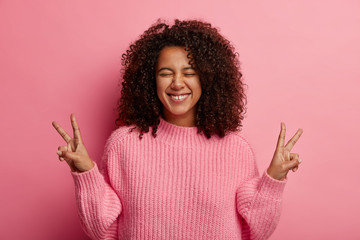 Playful carefree woman with Afro hairstyle, shows peace or victory sign, enjoys life, wears...