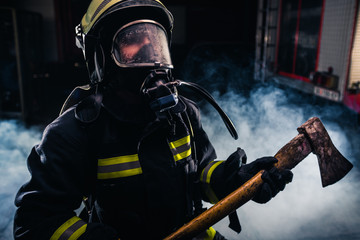 Naklejka premium Portrait of a female firefighter while holding an axe and wearing an oxygen mask indoors surrounded by smoke.