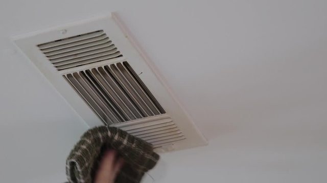 Dusting HVAC ceiling air vent with dust cloth. Cleaning overhead home heat and air conditioning ventilation duct.