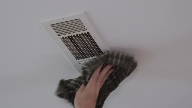 Dusting HVAC ceiling air vent with dust cloth. Cleaning overhead home heat and air conditioning ventilation duct.