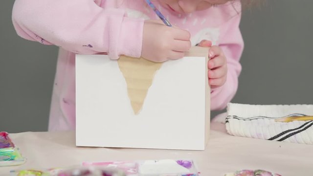 Little girl painting a white unicorn with acrylic paint on a wooden box.