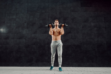 Handsome muscular caucasian blond shirtless man lifting dumbbells while standing in front of dark background.