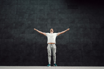 Powerful caucasian handsome fit bearded blond sportsman in tracksuit and in t-shirt holding hands in the air and looking up while standing in front of dark background.