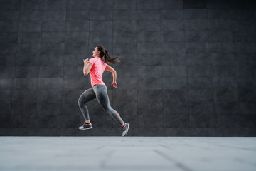 Obraz na płótnie Canvas Side view of fit attractive Caucasian woman in sportswear and with ponytail running outdoors. In background is dark wall.