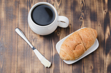 French breakfast. White mug with black coffee and croissant with butter knife on wooden table. Top view. Coffee break.