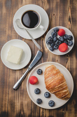 Fototapeta na wymiar Coffee cup, croissant with berries in white bowl and butter knife on wooden table. Top view. Healthy breakfast with fresh berries