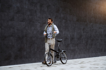 Naklejka premium Full length of cheerful handsome caucasian fashionable man listening music and pushing bicycle. In background is gray wall.