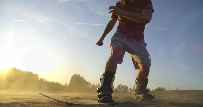 SLOW MOTION hipster man athlete in shorts tries doing tricks with a Board on sand dunes. Sandboard, snowboard, sunset, desert, flare