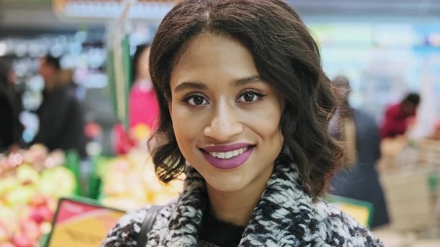 Beautiful woman looking at camera and smiling with grocery at background. Beautiful african american woman in supermarket. Close-up portrait in 4K, UHD