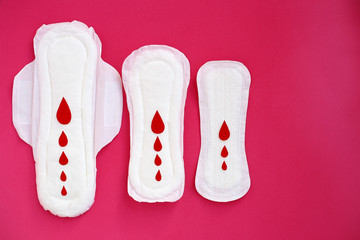 White woman cotton menstruation pads of different sizes with wings with red paper drops on pink background.