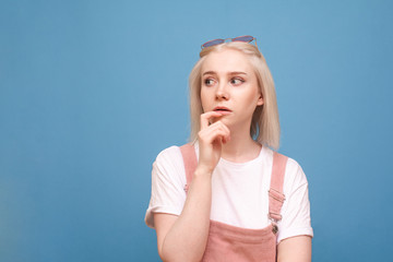 Beautiful blonde girl in cute clothes thoughtfully looking side on a blue background, wearing sunglasses white t-shirt and sunglasses.Thoughtful teenage girl looks out to the side of an empty space