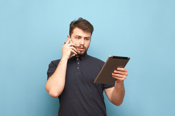 Busy man calls on the phone and looks at the screen of a tablet on a blue background wearing a beard and a dark t-shirt. Business man uses a tablet on a smartphone, isolated.
