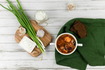 red borsch, a traditional national dish of Ukrainian cuisine. on the stock photo is served in a white pottery with cream, green onions and garlic on a light wooden background. green textile. flat lay