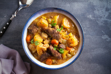 Beef meat stewed with potatoes, carrots and spices in bowl on dark gray background. Overhead horizontal image