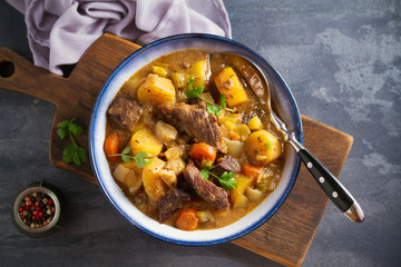 Beef meat stewed with potatoes, carrots and spices in bowl on dark gray background. Overhead horizontal image