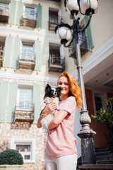 The theme is the friendship of man and animal. Beautiful young red hair Caucasian woman holding a pet dog Chihuahua breed near a house building in the summer in sunny weather