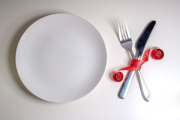 Losing weight. A fork and knife are wrapped in a red measuring tape next to a white plate with a white background