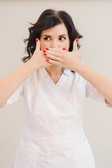 Naklejka premium A young girl in a medical coat shows with both hands a sign of silence. A mature brunette model in a cosmetology white suit covered her mouth with her hands on a beige background in the studio