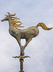 Melbourne, Australia - November 16, 2009: Small greenish bronze statue of horse with golden manes and tail seems to gallop and stands on a pole against light blue sky.