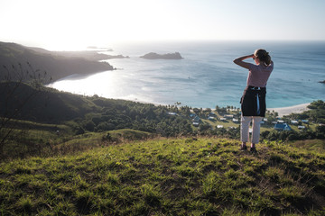 Girl standing on the top of the green mountain, admiring view of a small village with ocean in...
