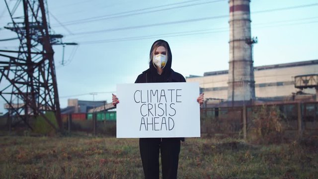 Climate Crisis Ahead. Worried female activist standing near working factory holding poster in breathe protection mask looking at camera. Nature protection. Global warming potential. Air pollution.