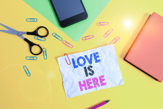 Text sign showing Love Is Here. Business photo text Roanalysistic feeling Lovely emotion Positive Expression Care Joy Paper sheets pencil clips smartphone scissors notebook colored background