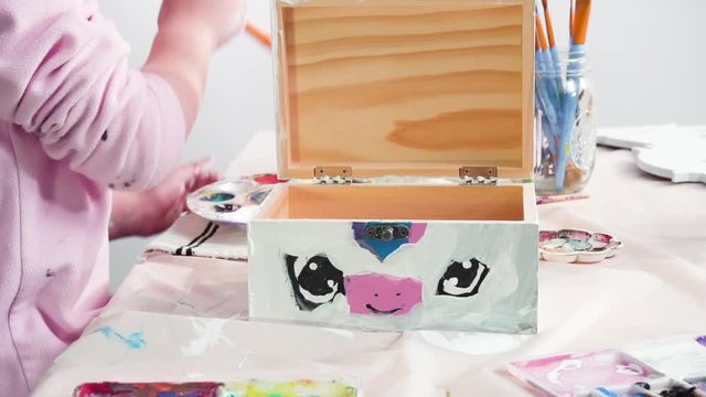 Time lapse. Little girl painting a white unicorn with acrylic paint on a wooden box.
