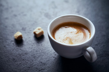 Cup of coffee and two brown sugar cubes on dark stone background. Close up.