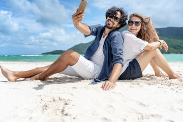 Fototapeta premium Beautiful young couple joyfully sitting on the white sand by the sea and takes a selfie on a smartphone. Honeymoon Travel and Vacation on the Tropical Ocean