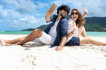Beautiful young couple joyfully sitting on the white sand by the sea and takes a selfie on a smartphone. Honeymoon Travel and Vacation on the Tropical Ocean