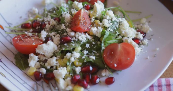 Fresh made arugula salad with cherry tomatoes, soft cheese, sweetcorn kernel and pomegranate seed on plate on rotating stand, close up