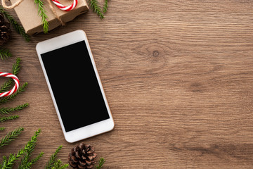 Smartphone with blank screen is in the middle of wood table with christmas decorations. Top view with copy space, flat lay.