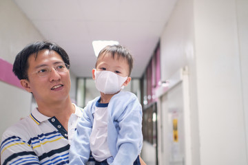 Father holding cute little Asian 3 years old toddler boy child wearing protective medical mask, parent and sick kid, Dad and son waiting to see the doctor at hospital with copy space