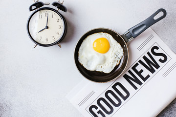  Breakfast time. Fried eggs in a pan with an alarm clock and a newspaper on a light gray table with a textured background.