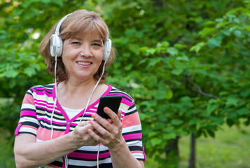 Senior beautiful woman listens to music in headphones with phone in her hands in summer park. Hiking, active and healthy lifestyle