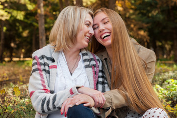 Fototapeta premium Happy adult daughter hugs her mom sitting in summer park, they are laughing. Tender and caring relationships of children and parents
