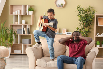 African-American man covering ears while his little son is playing guitar at home