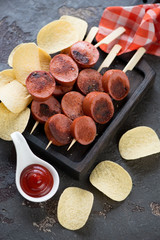 Black wooden serving tray with slices of roasted sausages on skewers and chips, vertical shot over brown stone background