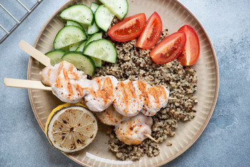 Beige plate with chicken skewers, quinoa and fresh vegetables, flatlay over light-blue stone background, horizontal shot