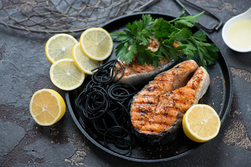 Grilled salmon steaks with squid-ink spaghetti, lemon and parsley on a round metal tray, studio shot over brown stone surface