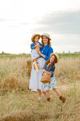 Naklejka na ściany i meble Beautiful happy mother in a straw hat with two little cute daughters standing in the middle of a bread field on a sunny summer day and holding buns in their hands