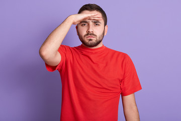Fototapeta na wymiar Close up portrait of serious handsome bearded man looking into distance, holding hand at forehead, posing isolated against blue background, attractive guy wearing red casual t shirt. People concept