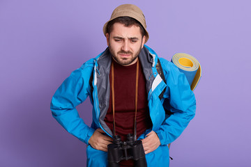 Studio shot of young handsome man standing isolated overblue background with hands on stomach because indigestion, traveler with mat and binoculars,wearing jacket and hat. Healthy care concept.