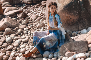 beautiful young woman in elegant stylish dress on stone beach at sunset