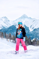 Fototapeta na wymiar young happy girl stands on a snowboard in bright colored clothes on a background of snowy mountains