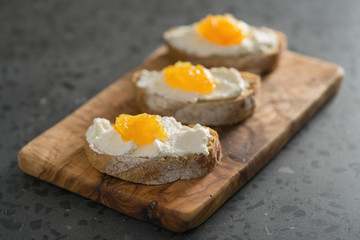 Three ciabatta slices with ricotta and orange marmalade on olive wood board on terrazzo surface