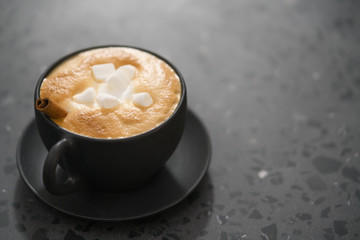 cappuccino with marshmallow and cinnamon in black cup on terrazzo surface with copy space