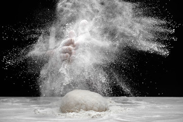 White flour flies in air on black background, pastry chef claps hands and prepares yeast dough for...
